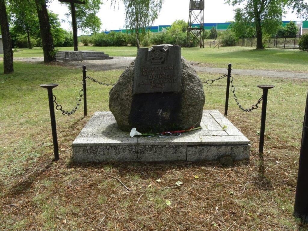 Kamienny pomnik z tablicą pamiątkową stoi na brukowanej platformie, otoczony czterema słupami połączonymi łańcuchami. Upamiętnia rocznicę rozstrzelania gen. Henryka Kowalówki i znajduje się w trawiastym parku z drzewami i pobliską wieżą strażniczą.