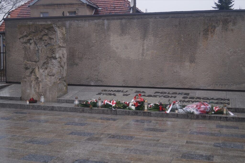 Ściana pamiątkowa z wygrawerowanym tekstem i płaskorzeźbą, na której znajdują się kwiaty i wieńce złożone u podstawy na mokrym kamiennym chodniku. Ponura scena oddała hołd ofiarom Niemieckich Nazistowskich Obozów Koncentracyjnych w Narodowym Dniu Pamięci.