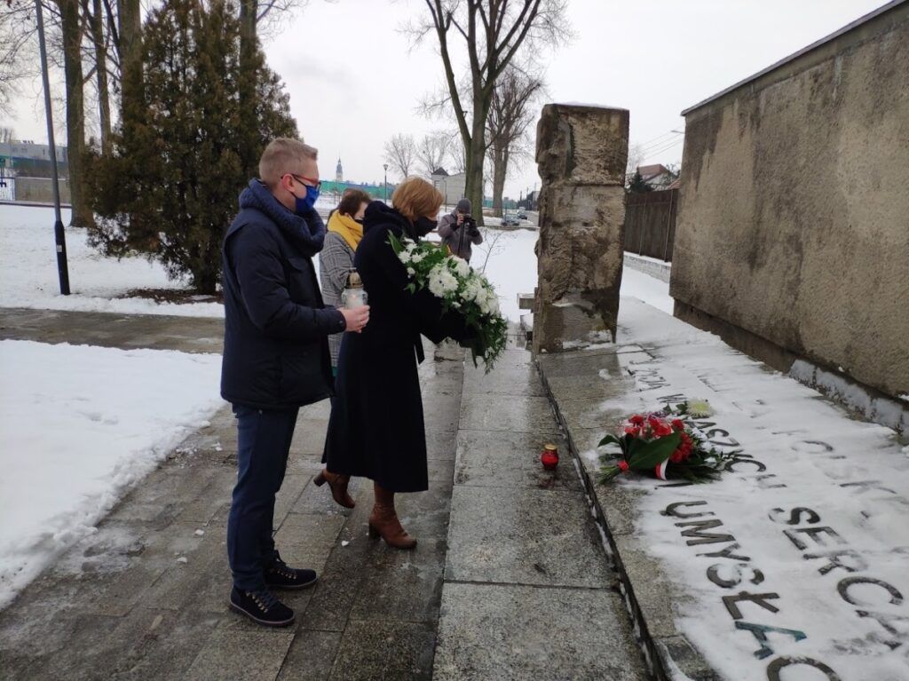 Dwie osoby stoją przed pokrytym śniegiem pomnikiem, trzymając wieńce z kwiatów, oddając hołd rocznicy obozu karno-śledczego. Kolejny wieniec i świeca spoczywają na ziemi, gdy oddają hołd w zimny zimowy dzień.