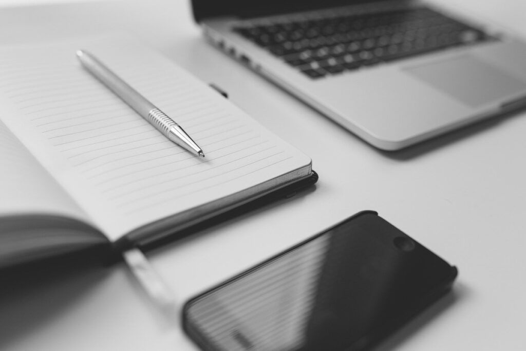 Srebrny długopis leży na otwartym notesie w linie obok smartfona i częściowo widocznego laptopa na białym biurku. Scena, w czerni i bieli, przywołuje osobiste historie czekające na zapisanie lub opowiedzenie podczas webinarium.