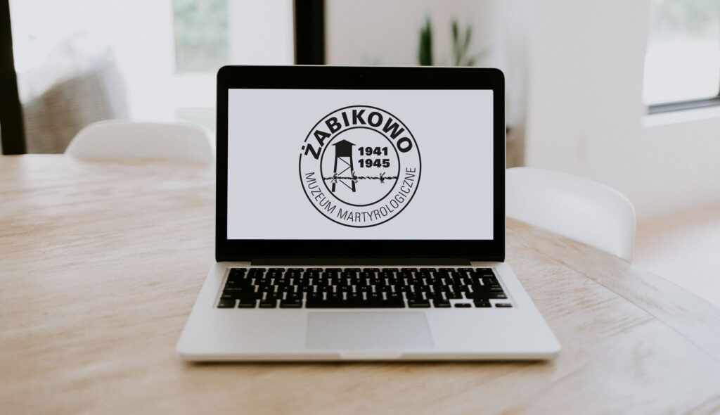 Na drewnianym stole znajduje się laptop z logo Muzeum Martyrologii w Żabikowie, na którym widnieje tekst oraz lata 1941–1945 ze stylizowanym wzorem drutu kolczastego, upamiętniającym Pracę przymusową Polaków w Kraju Warty.