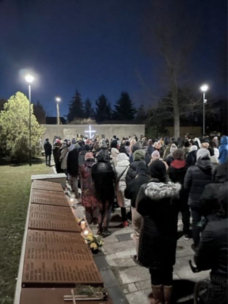 Duża grupa ludzi, ubranych w ciepłe ubrania, zbiera się nocą na zewnątrz przy ścianie pamięci obóz karno-śledczy Żabikowo z tablicami pamiątkowymi i zapalonym krzyżem w tle podczas Drogi Krzyżowej w 2023 r. W pobliżu tablic umieszczono świece i kwiaty.