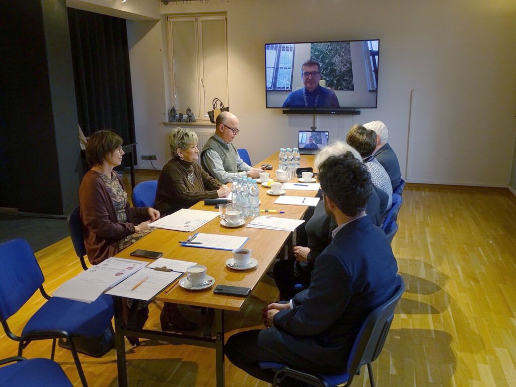 Członkowie Rady Muzeum siedzą przy stole konferencyjnym z dokumentami i napojami, a dwa inne elementy pojawiają się w Internecie, pojawiając się na zauważalnym w nowoczesnym, jasnym sali.