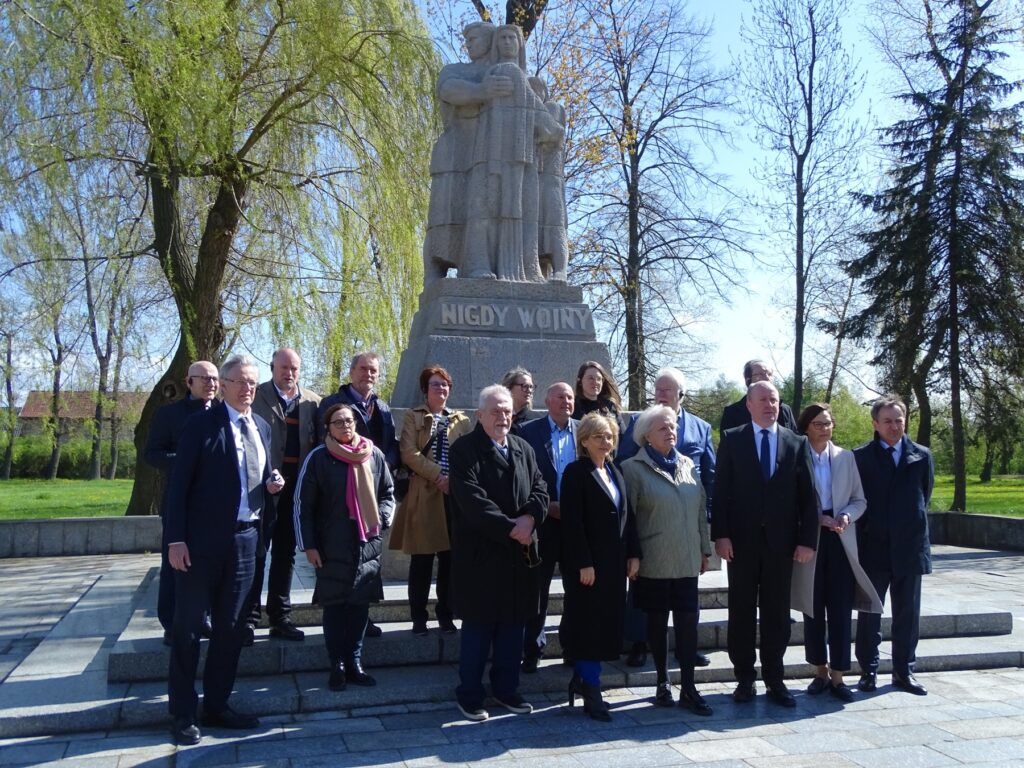 Grupa osób, w tym Delegacja Parlamentu Hesji, pozuje przed kamiennym pomnikiem z napisem NIGDY WOJNY w Muzeum Martyrologicznym w Żabikowie w słoneczny dzień, na tle drzew i błękitnego nieba.