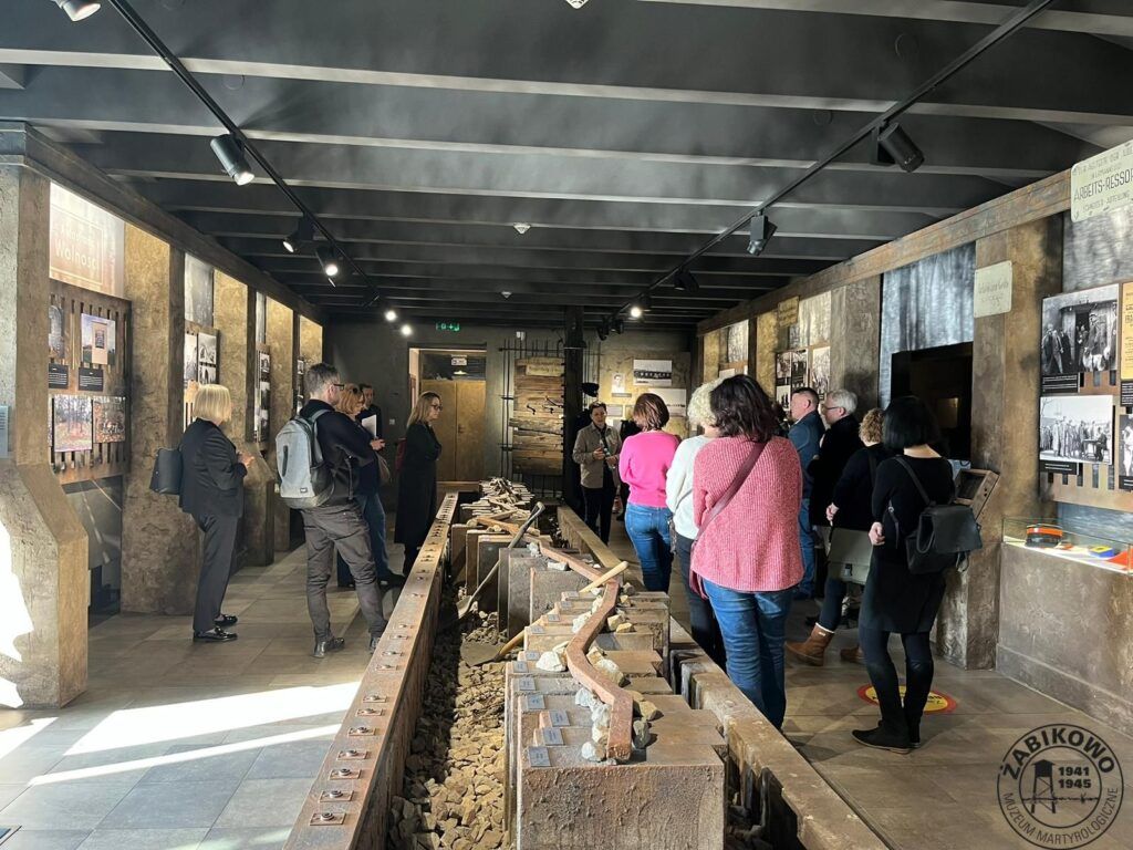 Grupa osób bierze udział w pobyt studyjny, zwiedzając wystawę muzeum historycznego z maszynami przemysłowymi, torami kolejowymi i informacyjnymi ekspozycjami ściennymi. Pomieszczenie szczyci się kamienno-metalowym wzornictwem przemysłowym.