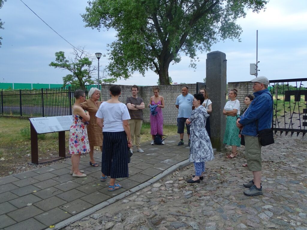 Podczas wizyty studyjnej w Muzeum KL Plaszow grupa stoi na zewnątrz w pobliżu znaku informacyjnego i bramy, słuchając kobiety. W tle pod zachmurzonym niebem widać drzewa, bruk i ogrodzenie.