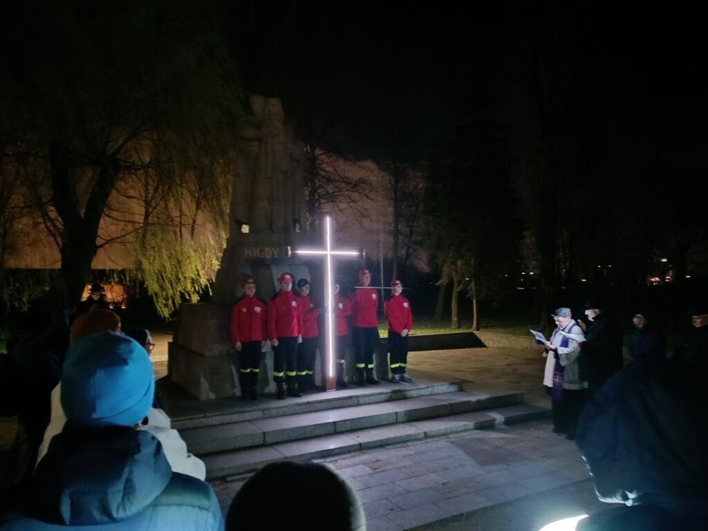 Grupa ludzi zbiera się nocą wokół oświetlonego krzyża przed pomnikiem Drogi Krzyżowej. Kilka osób w czerwonych kurtkach stoi przy krzyżu, podczas gdy inni oglądają lub czytają z gazet w pobliżu. W tle widać drzewa i posąg.