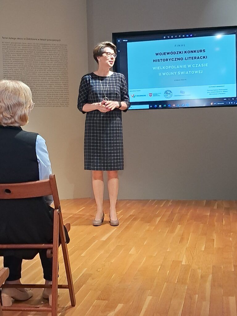 Kobieta w kraciastej sukience stoi i przemawia przed ekranem wyświetlającym prezentację z wydarzenia Konkurs Historyczno-Literacki, skupionego na II wojnie światowej. Dwie osoby siedzą na krzesłach naprzeciwko niej w pokoju z drewnianą podłogą.