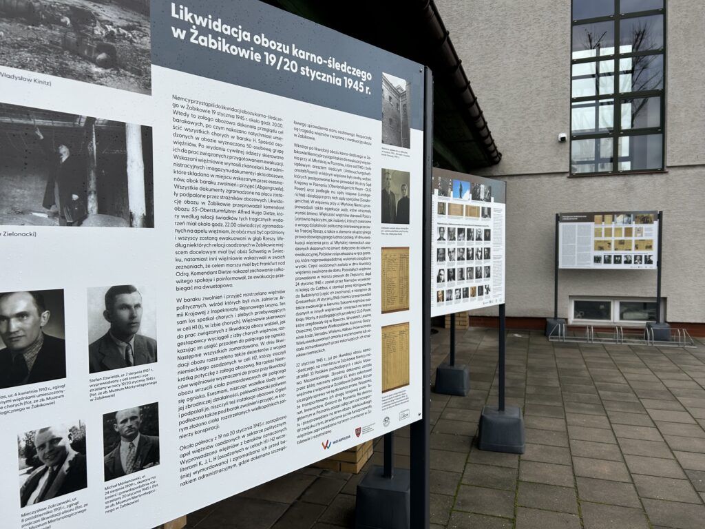 Wystawa plenerowa z tablicami informacyjnymi w języku polskim prezentuje teksty, zdjęcia i dokumenty o ewakuacji obozu karno-śledzonego Żabikowo. Tablice są na stojakach w pobliżu nowoczesnego budynku.