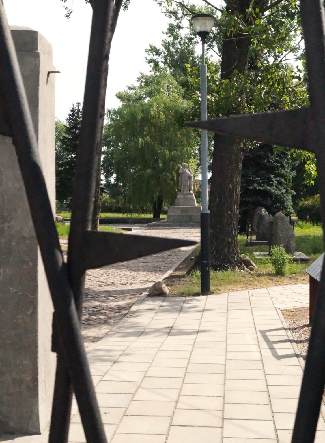 Widok przez metalową bramę z kolcami ukazuje brukowaną ścieżkę, drzewa, latarnię i odległy pomnik oznaczający miejsce dawnego obozu karno-śledczego Żabikowo. Na pomniku widnieje napis "Nigdy wojny".