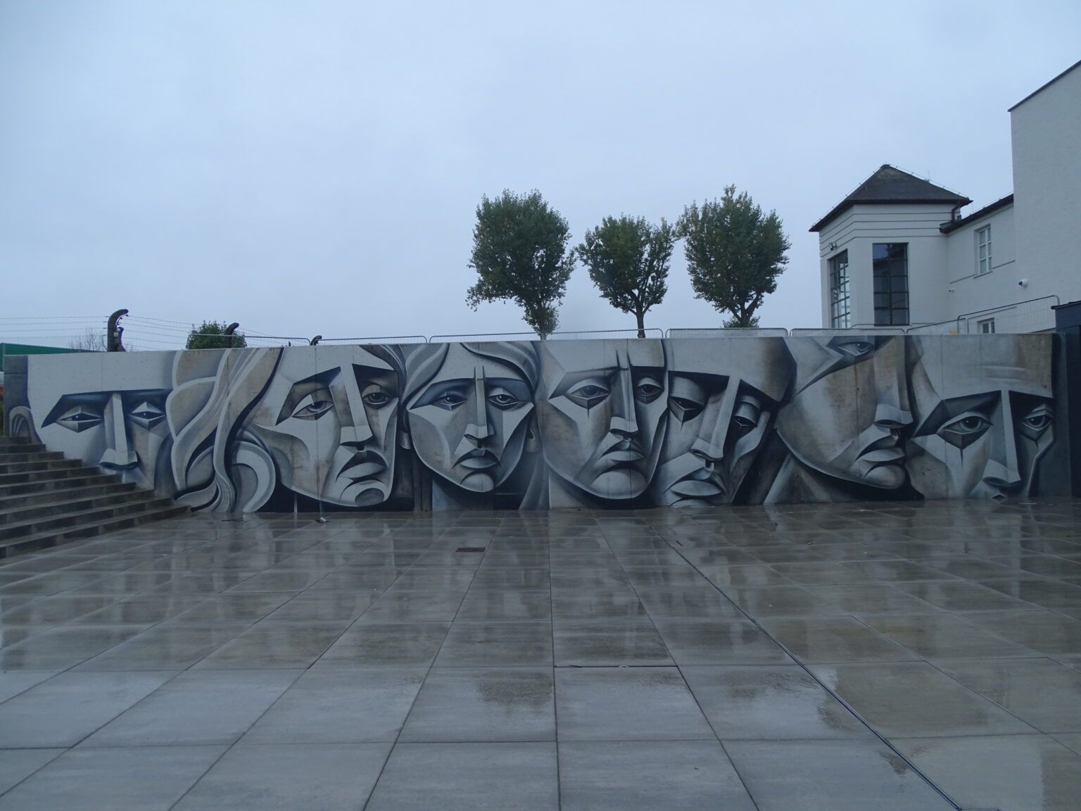 Mural na szarej ścianie przedstawia kilka dużych, ponurych ludzkich twarzy w odcieniach szarości. Podłoże jest mokre, a w tle widoczne są trzy drzewa i nowoczesne budynki pod zachmurzonym niebem, co sugeruje, że ukończenie prac nastąpiło niedawno.