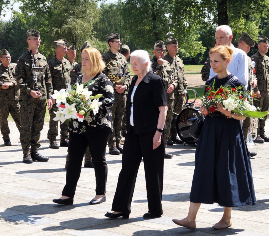 Trzy kobiety niosą wiązanki kwiatów, idąc po terenie dawnego obozu przy pomniku gen. Kowalówka w Muzeum w Żabikowie.
