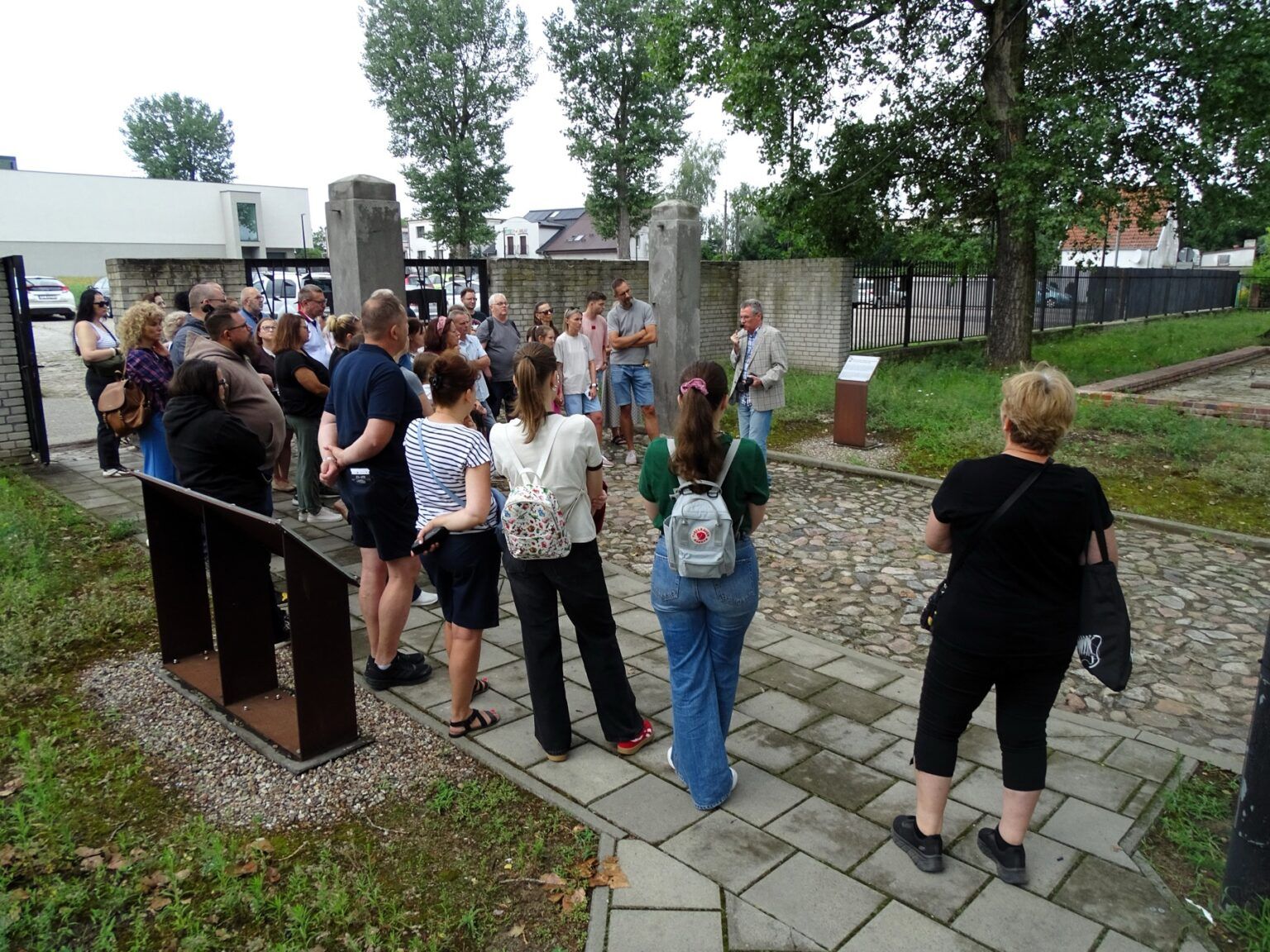 Grupa osób słucha przewodnika podczas zwiedzania terenu dawnego obozu w Muzeum Martyrologicznym w Żabikowie, stojąc na kamiennej ścieżce przy ogrodzeniu, niedaleko ekspozycji muzealnej i pomników upamiętniających ofiary obozu.