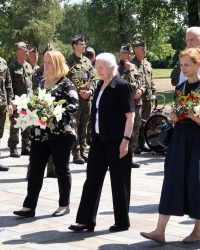 Trzy kobiety niosą wiązanki kwiatów, idąc po terenie dawnego obozu przy pomniku gen. Kowalówka w Muzeum w Żabikowie.