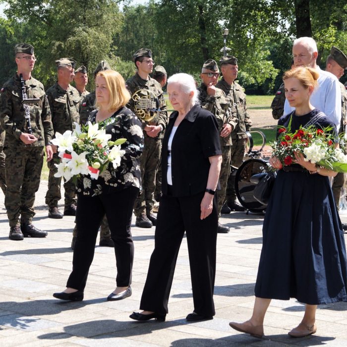 Trzy kobiety niosą wiązanki kwiatów, idąc po terenie dawnego obozu przy pomniku gen. Kowalówka w Muzeum w Żabikowie.