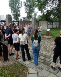 Grupa osób słucha przewodnika podczas zwiedzania terenu dawnego obozu w Muzeum Martyrologicznym w Żabikowie, stojąc na kamiennej ścieżce przy ogrodzeniu, niedaleko ekspozycji muzealnej i pomników upamiętniających ofiary obozu.