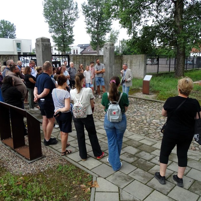 Grupa osób słucha przewodnika podczas zwiedzania terenu dawnego obozu w Muzeum Martyrologicznym w Żabikowie, stojąc na kamiennej ścieżce przy ogrodzeniu, niedaleko ekspozycji muzealnej i pomników upamiętniających ofiary obozu.