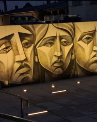 Fragment muralu w Muzeum Martyrologii w Żabikowie z trzema stylizowanymi twarzami, oświetlony nocą przed budynkiem muzeum.