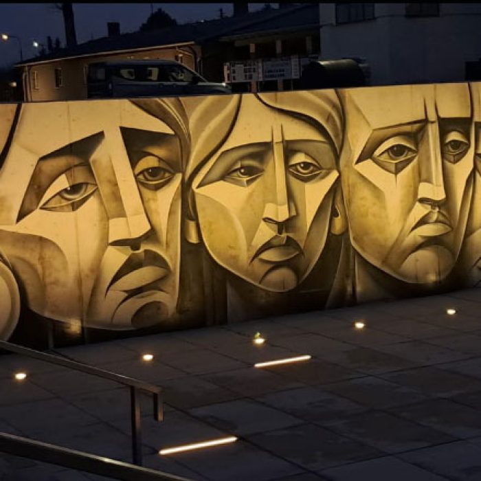 Fragment muralu w Muzeum Martyrologii w Żabikowie z trzema stylizowanymi twarzami, oświetlony nocą przed budynkiem muzeum.