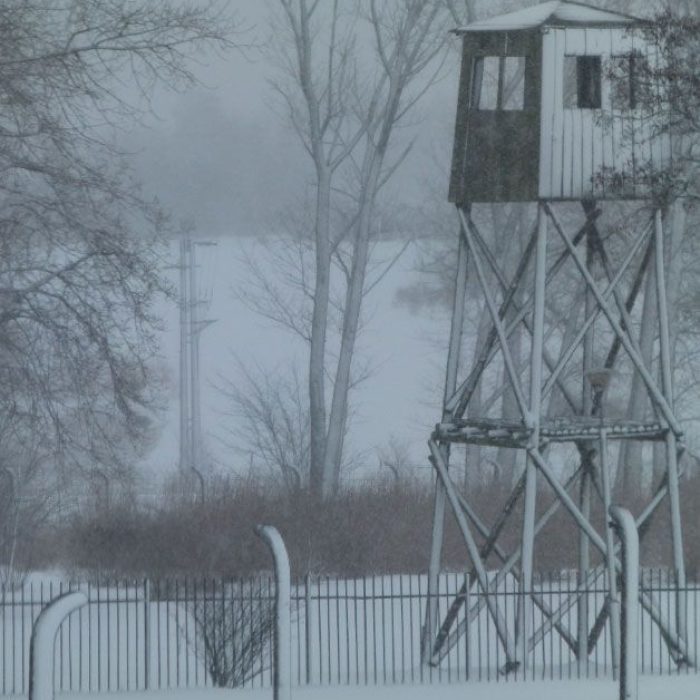 Wysoka, metalowa wieża strażnicza stoi za ogrodzeniem z drutu kolczastego w zaśnieżonym krajobrazie Żabikowa, przywołując mrożącą krew w żyłach historię związaną z ewakuacją Obozu Karno-Śledczego pośród nagich drzew i gęstej mgły.